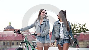 Portrait of two young attractive urban women walking with a bike and skateboarding on the street in sunny summer day.