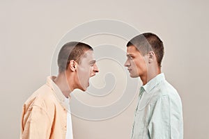 Portrait of two young caucasian men, twin brothers arguing, shouting while standing face to face isolated over beige