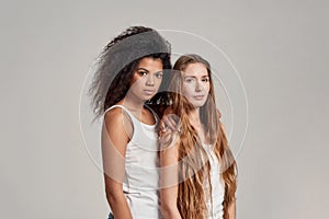 Portrait of two young diverse women wearing white shirts looking at camera while posing together isolated over grey