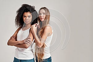 Portrait of two young diverse women wearing white shirts looking at camera while posing together isolated over grey