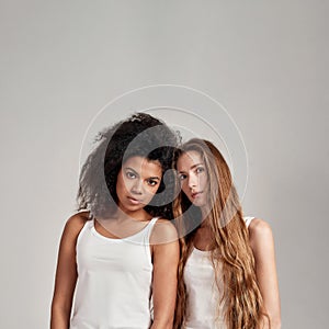 Portrait of two young diverse women wearing white shirts looking at camera while standing together isolated over grey