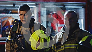 Portrait of two young firefighters on the rain in front of fire engine in full uniform. Fire dril