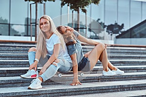 Portrait of a two young hipster girls sitting together on skateboard at steps on a background of the skyscraper.