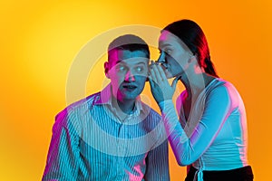 Portrait of two young people, woman whispering on man's ear, talking news isolated over orange background in neon