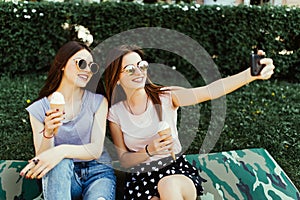 Portrait of two young pretty women standing together eating ice cream and taking selfie photo on camera in summer street.