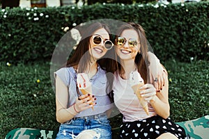 Portrait of two young women standing together eating ice cream hugging sitting on the grass in city street.