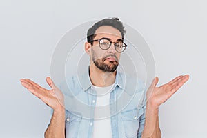 Portrait of uncertain young stylish stubble man with trendy round glasses wears demin blue shirt, shrugs shoulders being puzzled