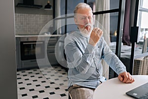 Portrait of unhappy aged gray-haired man eating unappetizing burger at kitchen home. Hungry older bearded disappointed