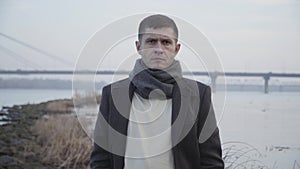 Portrait of unhappy Caucasian man posing at the background of river and city bridge. Upset young guy looking at camera