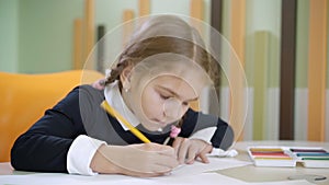 Portrait of unhappy girl drawing at desk and throwing pencil away. Cute little Caucasian schoolgirl having problems on