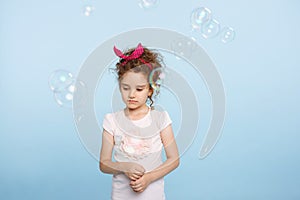 Portrait unhappy little girl, posing in studio, looking down over light blue background. Soap bubbles fly near kid.