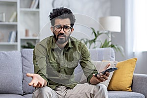 Portrait of unhappy man at home, indian man upset and disappointed sitting on sofa in living room and looking at camera