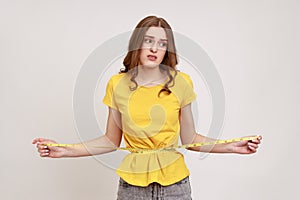 Portrait unhappy smiling woman in yellow T-shirt measuring her waist with tape measure and expressing sadness, looking away with