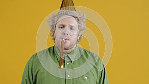 Portrait of unhappy young man taking off party hat blowing horn looking at camera