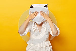 Portrait of unknown anonymous female wearing dress and paper cat mask posing  over yellow background, hiding her face,