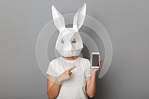 Portrait of unrecognizable woman wearing white t shirt and paper rabbit mask standing isolated over gray background, showing smart