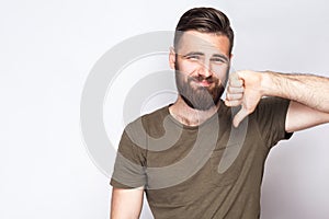 Portrait of unsatisfied bearded man with thumbs down and dark green t shirt against light gray background.