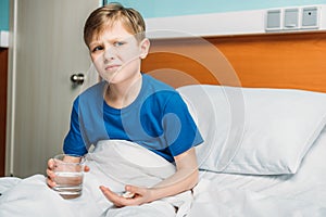 Portrait of unsatisfied boy holding glass of water and medicines in hospital bed