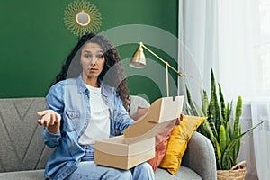 Portrait of unsatisfied female shopper in online store, Hispanic woman looking disappointed at camera sitting on sofa in