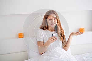 Portrait of unsatisfied girl sit bed under blanket hold smart phone shrug shoulder bright interior bedroom indoors