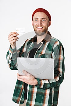 Portrait of unshaved man holding silver laptop and credit card, while standing isolated over white background