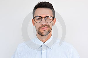 Portrait of unshaven handsome male frowns, being dissatisfied with something, blink with eye, wears round glasses and blue shirt,
