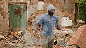 Portrait of upset adult african man standing in front of old ruined house after hurricane outdoors