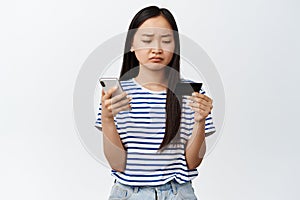 Portrait of upset asian girl frowning, looking at credit card and using mobile phone app, standing against white