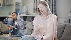 Portrait of upset beautiful slim woman sitting on couch looking back at blurred sad man. Stressed Caucasian girlfriend