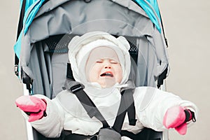 Portrait of upset crying Caucasian baby girl outside. Screaming child toddler in white warm clothes sitting lying in a stroller