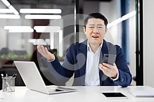 Portrait of upset japanese male holding personal smartphone while working at portable laptop in office environment