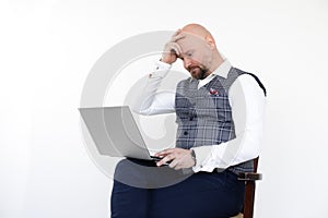 Portrait of upset middle-aged man in grey checkered vest, blue jeans, white shirt, sitting on chair, holding laptop.