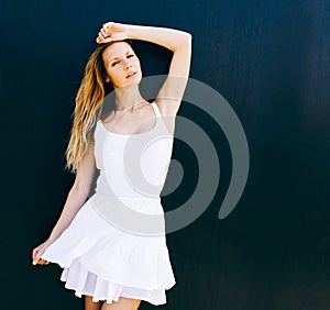 Portrait of very beautiful young blond woman in a short white dress posing on the street near a black wall. Sunny day. The wind bl