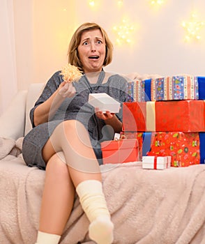 Portrait of a very surprised woman, sits on a couch and opens false gifts, holiday lights on a wall