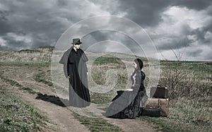 Portrait of a victorian lady in black sitting on the road with her luggage and gentleman standing nearby.