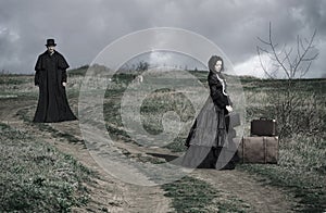 Portrait of a victorian lady in black sitting on the road with her luggage and gentleman walking down the road