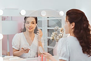 Portrait view of young adult asian woman looking at mirror, sitting in bright room and making make up. Beautiful and professional