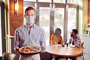 Portrait Of Waiter In Restaurant Wearing Mask Serving Pizza To Couple During Health Pandemic