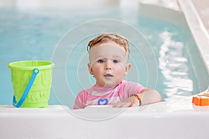 Portrait of white Caucasian baby girl playing with toys in water standing by swimming pool nosing inside, looking in camera, train photo
