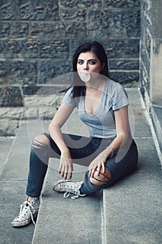 Portrait of white Caucasian beautiful young brunette girl with light blue green eyes in ripped jeans. Teenager blowing bubble gum