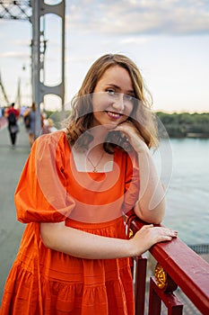 Portrait of white caucasian woman with red hair and blue eyes in an orange dress stands on the bridge and smiles to camera.