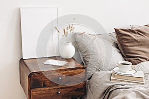 Portrait white frame mockup on retro wooden bedside table. Modern white ceramic vase, dry Lagurus ovatus grass. Cup of photo