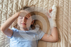 Portrait of a white girl with her arm in a cast due to an injury, in a blue T-shirt, lying on the bed in her room