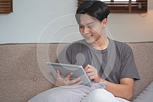 Portrait of a white man listening to music with a tablet in hand holding a pen on the sofa inside the house to relax on vacation