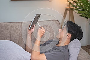 Portrait of a white man listening to music with a tablet in hand holding a pen on the sofa inside the house to relax on vacation