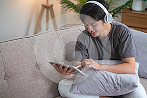 Portrait of a white man listening to music with a tablet in hand holding a pen on the sofa inside the house to relax on vacation