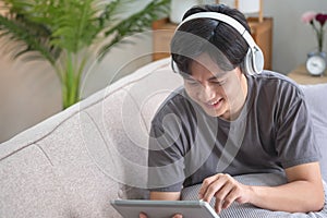 Portrait of a white man listening to music with a tablet in hand holding a pen on the sofa inside the house to relax on vacation