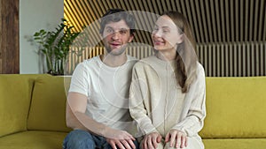 Portrait of a wife and husband waving their hands looking at the camera saying hello sitting on the sofa in the living