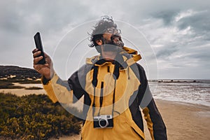 Portrait of wild photographer enjoying bad weather at the beach. Happy tourist with photo camera and mobile phone. Beach and ocean