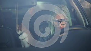 Portrait through window of young cheerful blonde driver girl with smartphone in hands and red cat inside car.